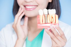 Dentist in white coat holding her hand to her jaw blurry in the background while holding up a model dental implant in the foreground